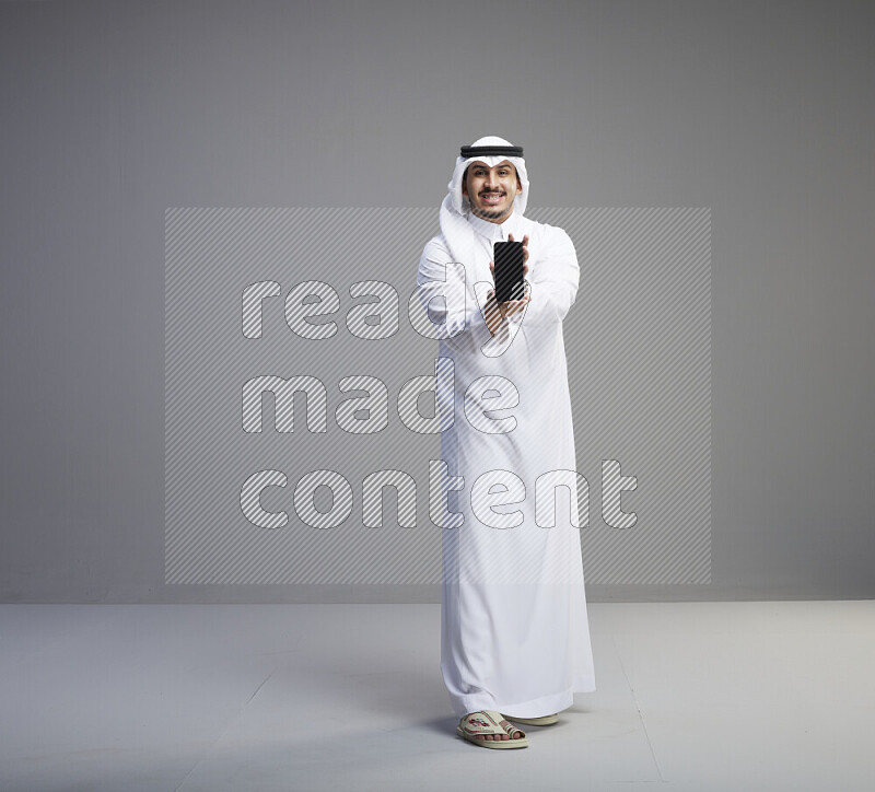 A Saudi man standing wearing thob and white shomag showing phone to camera on gray background