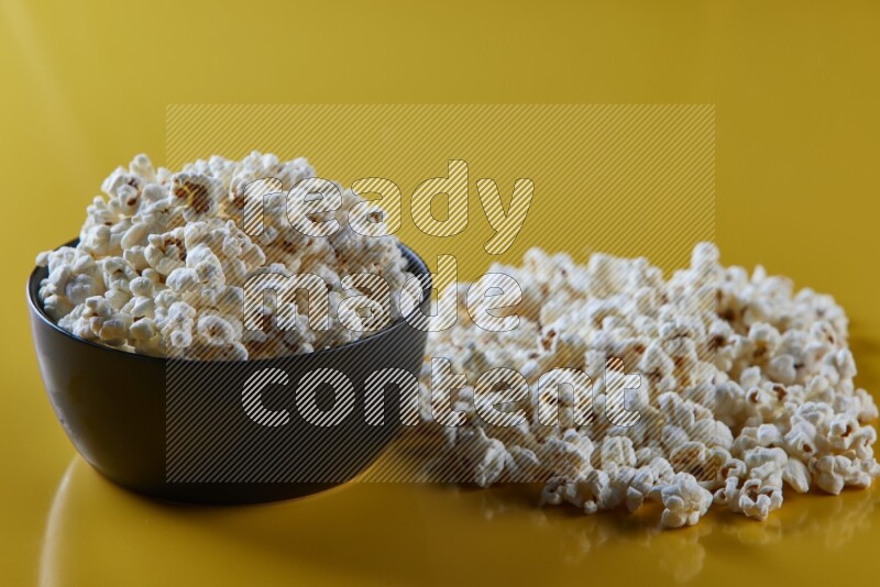 A black ceramic bowl full of popcorn with popcorn beside it on a yellow background in different angles