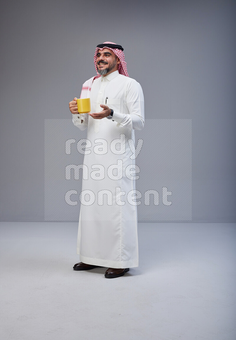 Saudi man Wearing Thob and red Shomag standing holding mug on Gray background