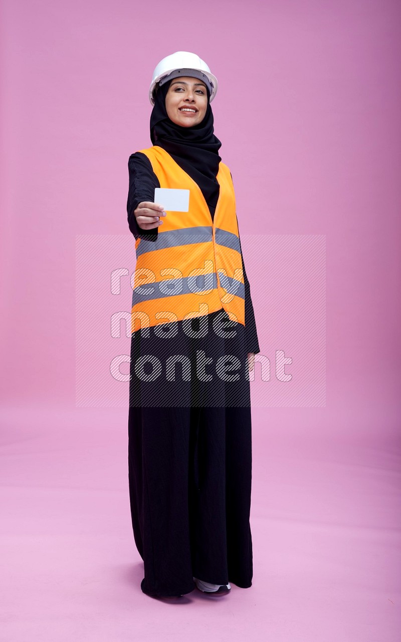 Saudi woman wearing Abaya with engineer vest and helmet standing holding ATM card on pink background