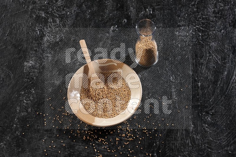 A beige pottery plate full of mustard seeds and a wooden spoon in it with a glass jar filled with the seeds on a textured black flooring