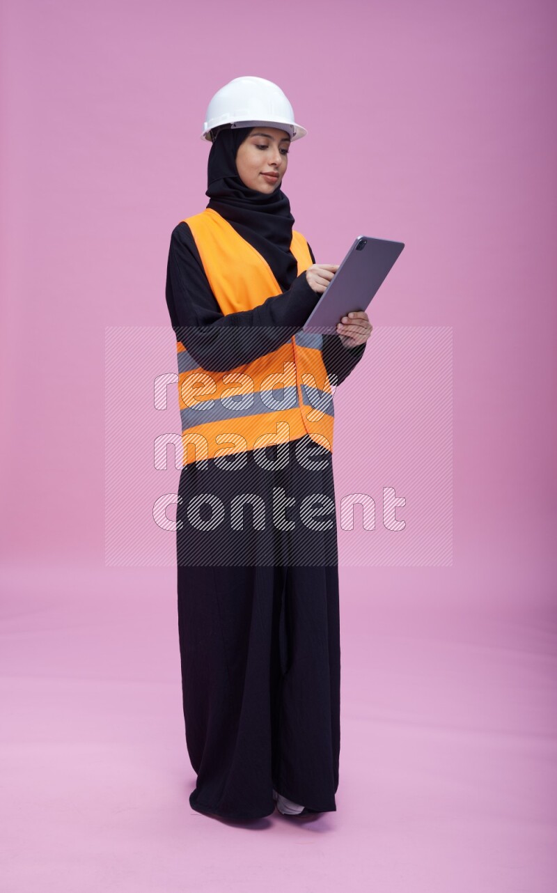 Saudi woman wearing Abaya with engineer vest and helmet standing working on tablet on pink background