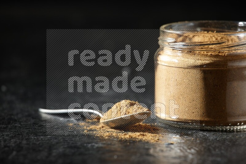 A glass jar and a metal spoon full of allspice powder on a textured black flooring