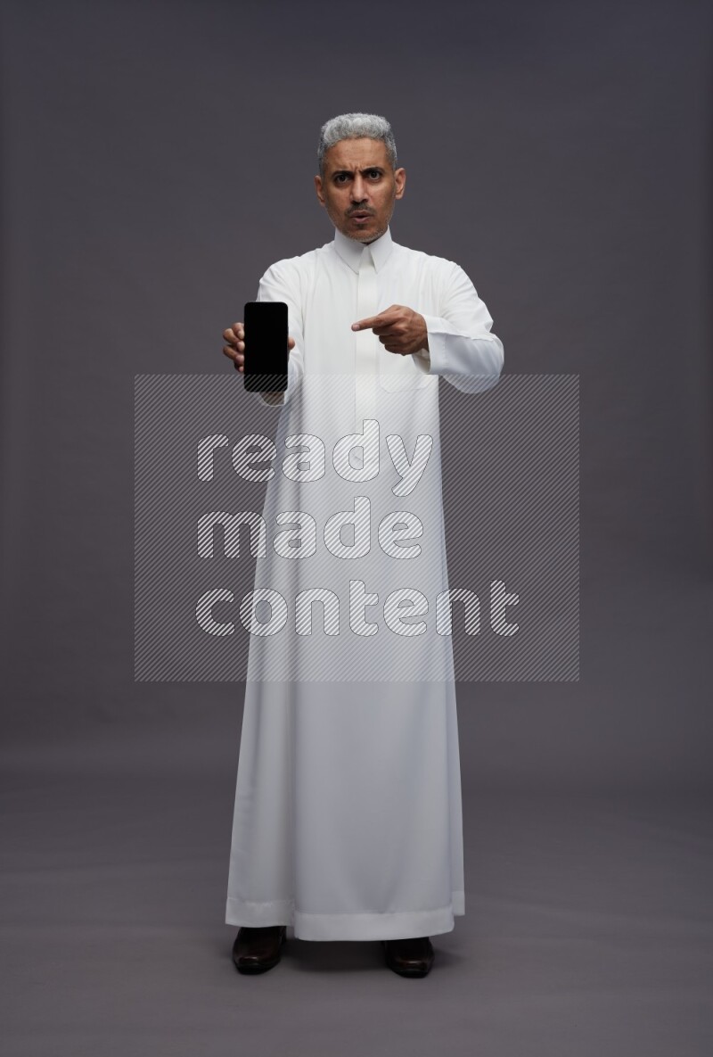 Saudi man wearing thob standing showing phone to camera on gray background