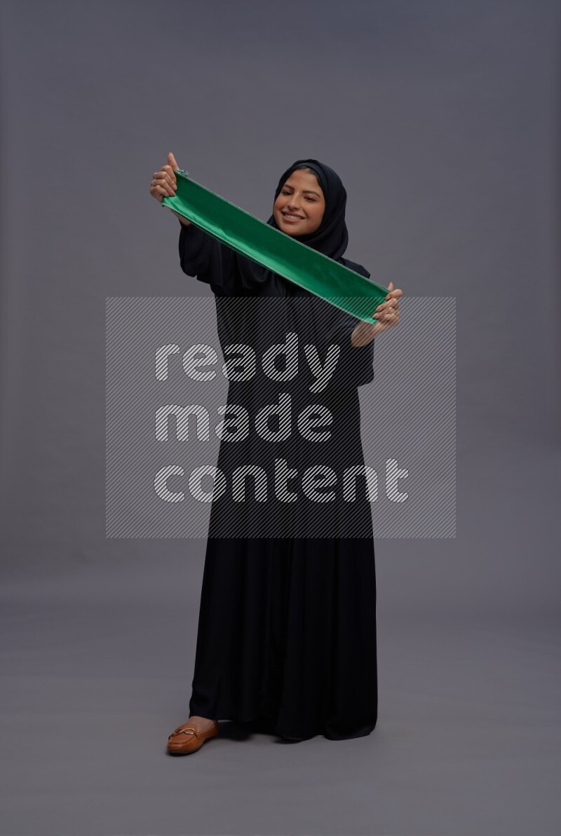 Saudi woman wearing Abaya standing holding Saudi flag on gray background