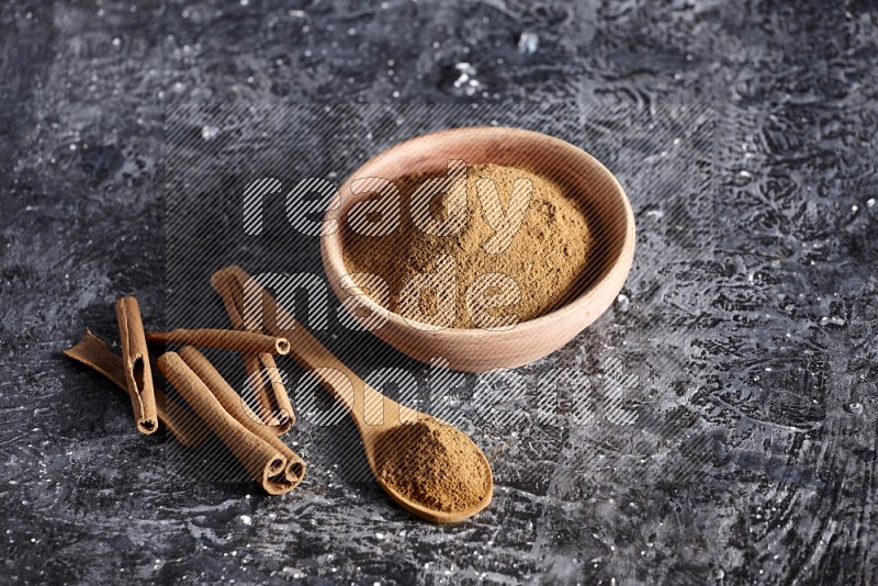 wooden bowl full of cinnamon powder and a wooden spoon full of it with cinnamon sticks on a textured black background