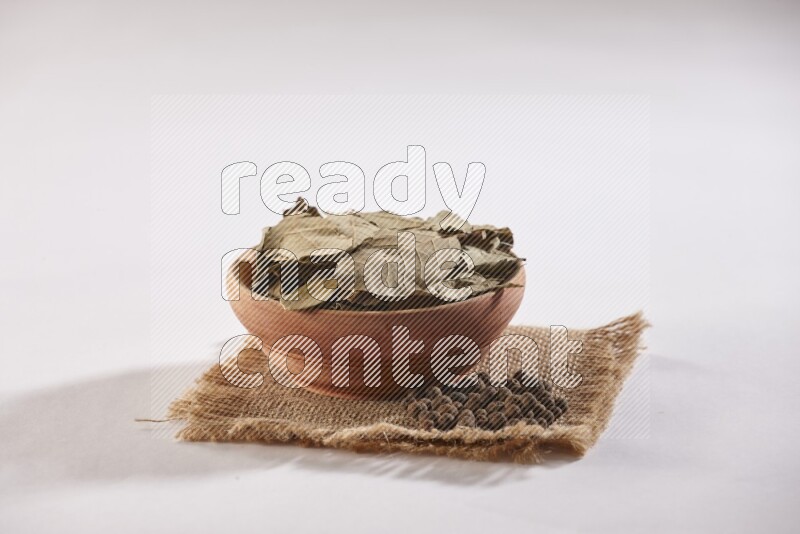 A wooden bowl filled with dried bay leaves on a piece of burlap with some of allspice berries on white flooring in different angles