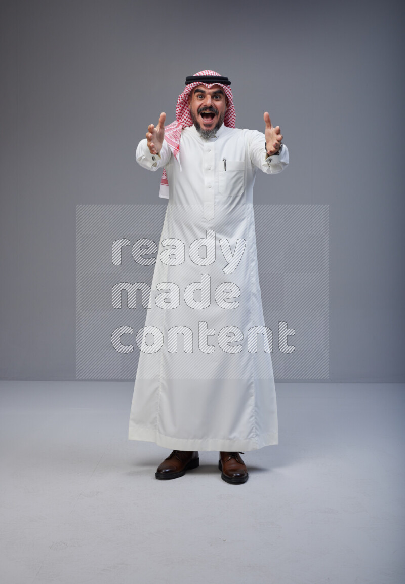 Saudi man Wearing Thob and red white Shomag standing interacting with the camera on Gray background