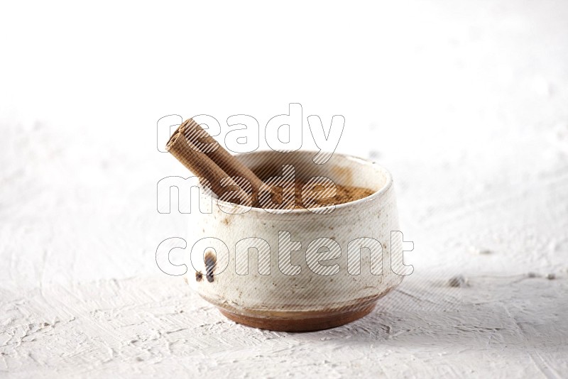 Ceramic beige bowl full of cinnamon powder with a cinnamon stick on a textured white background