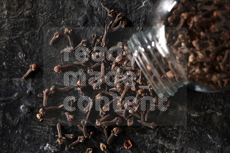 A flipped glass spice jar full of cloves and cloves came out of it on textured black flooring