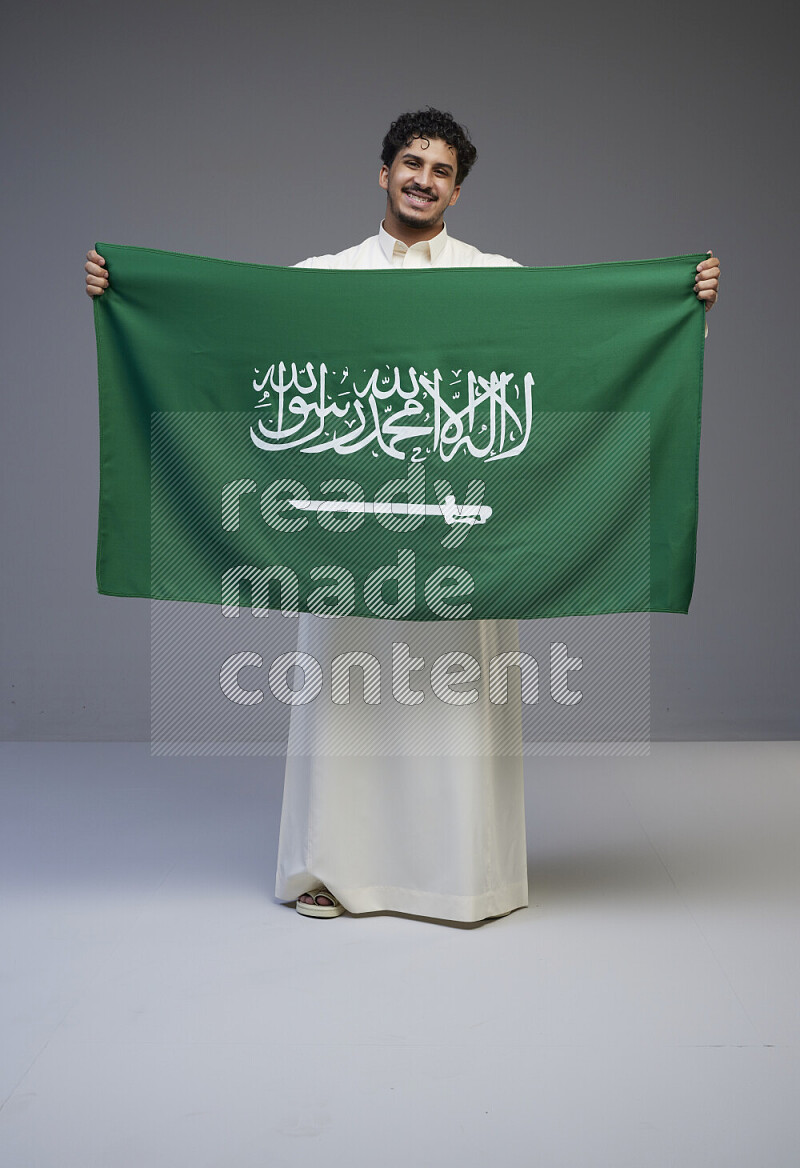 A Saudi man standing wearing thob holding big Saudi flag on gray background