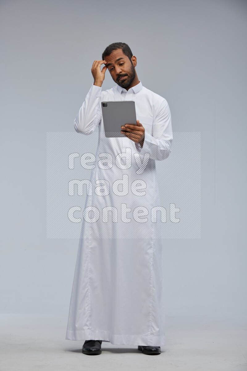 Saudi man wearing Thob standing working on tablet on Gray background