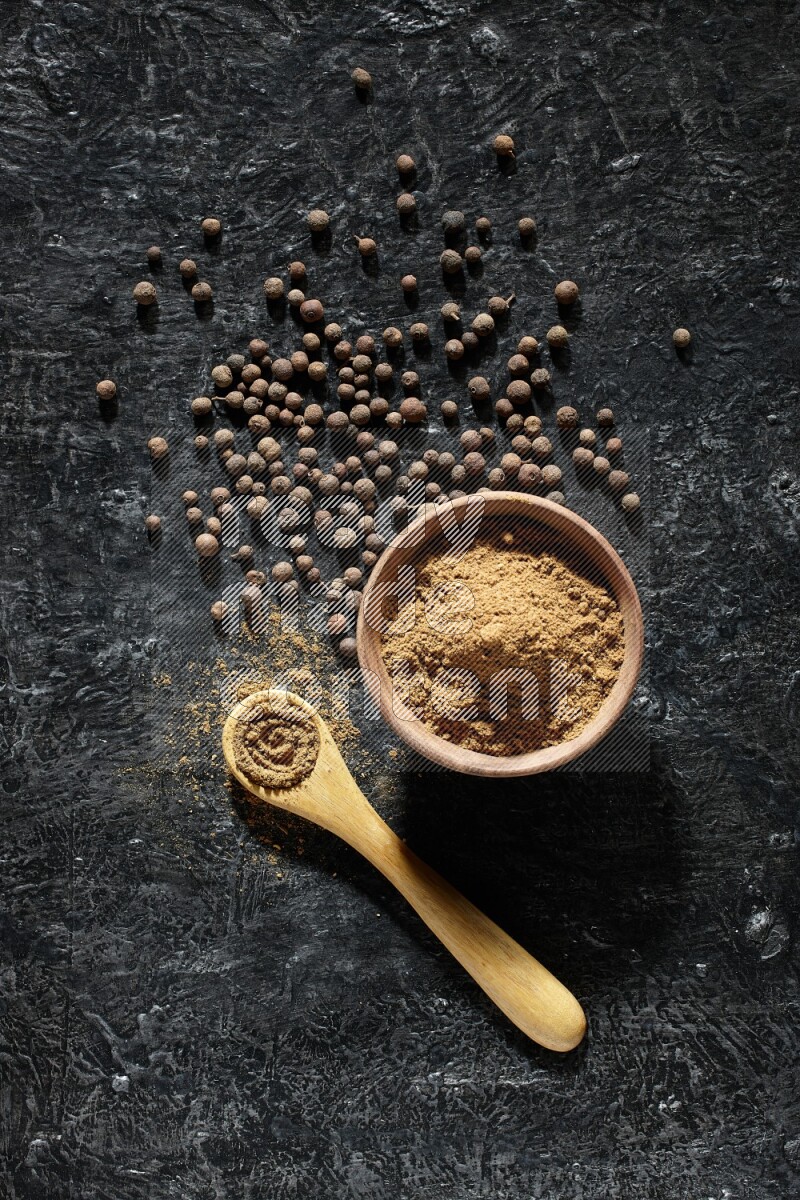 A wooden bowl and spoon full of allspice powder and whole balls spreaded on a black flooring
