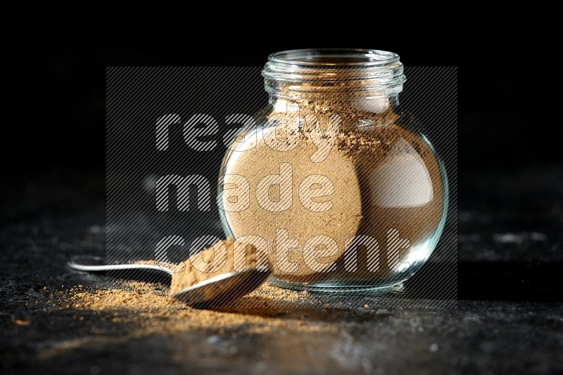 A glass spice jar and metal spoon full of allspice powder on a textured black flooring