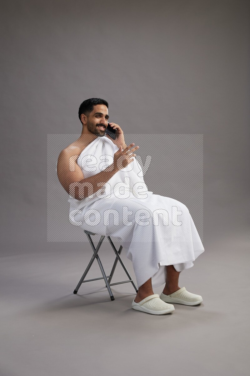 A man wearing Ehram sitting on chair talking on the phone on gray background