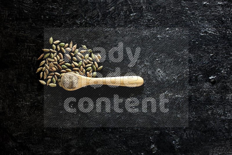 A wooden spoon full of cardamom powder and cardamom seeds next of it on textured black flooring