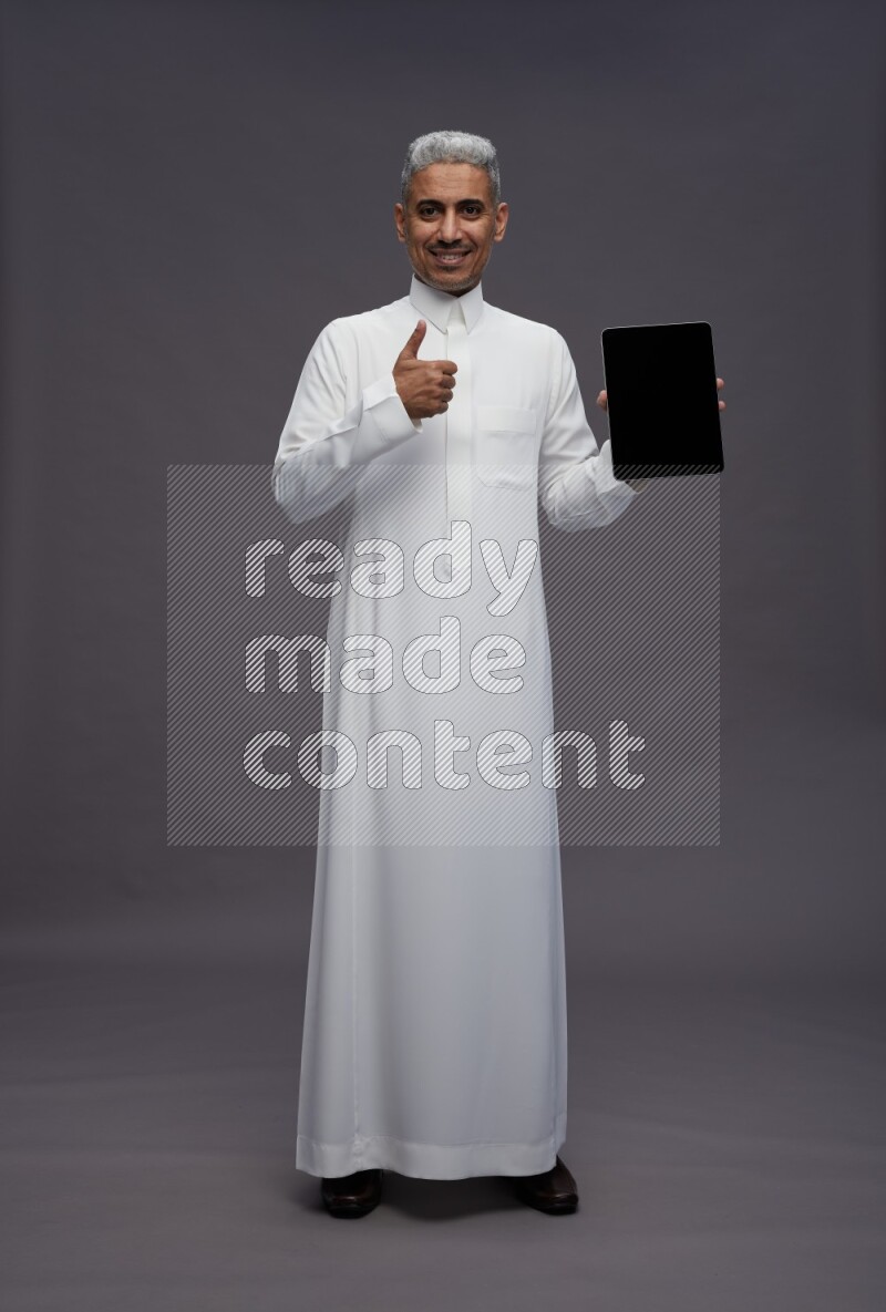 Saudi man wearing thob standing showing tablet to camera on gray background