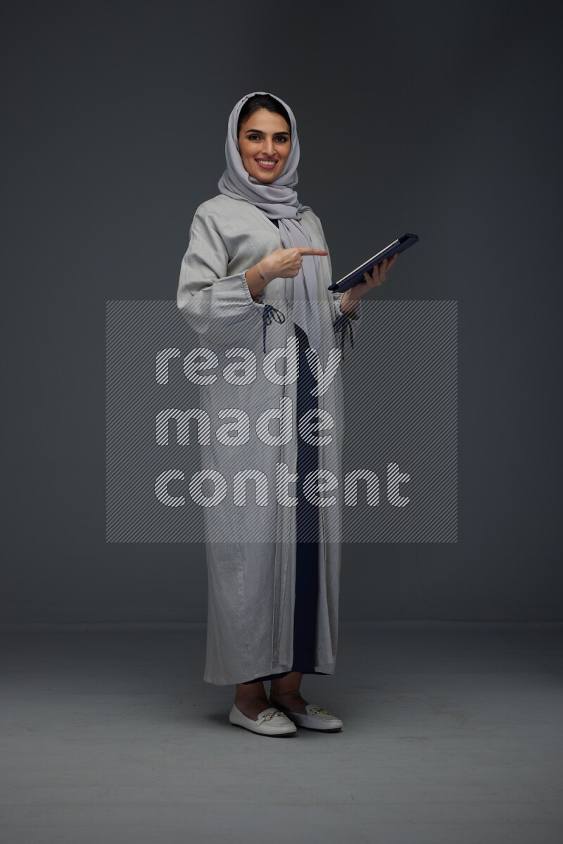 A Saudi woman wearing a light gray Abaya and head scarf standing holding a phone and pointing with the other hand eye level on a grey background