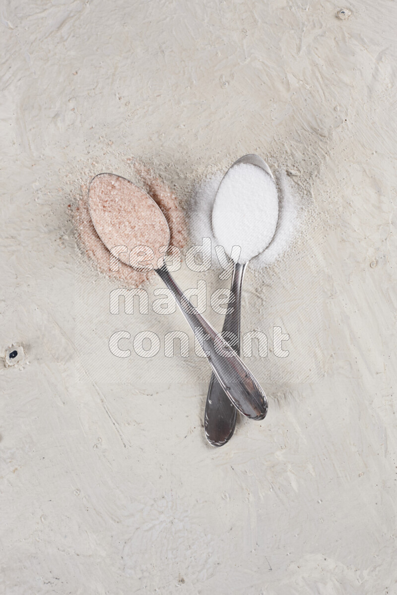 2 metal spoons filled with white salt and pink himalayan salt on white background