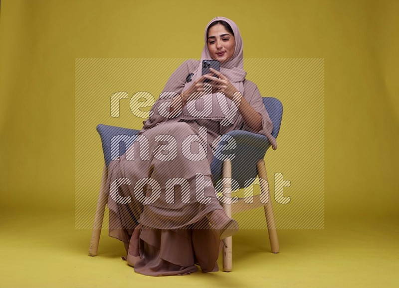 A Female Texting Sitting  on a Yellow Background wearing Brown Abaya with Hijab