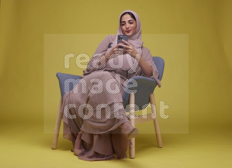A Female Texting Sitting  on a Yellow Background wearing Brown Abaya with Hijab