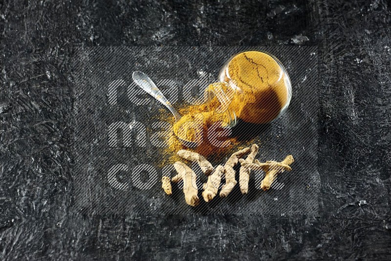 A flipped glass spice jar and metal spoon full of turmeric powder and powder spilled out of it with dried whole fingers on textured black flooring