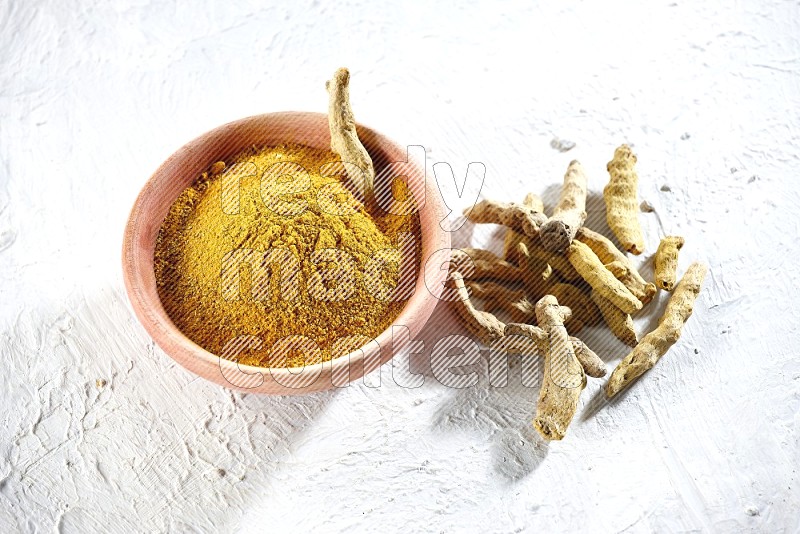 A wooden bowl full of turmeric powder and dried turmeric whole fingers beside it on textured white flooring