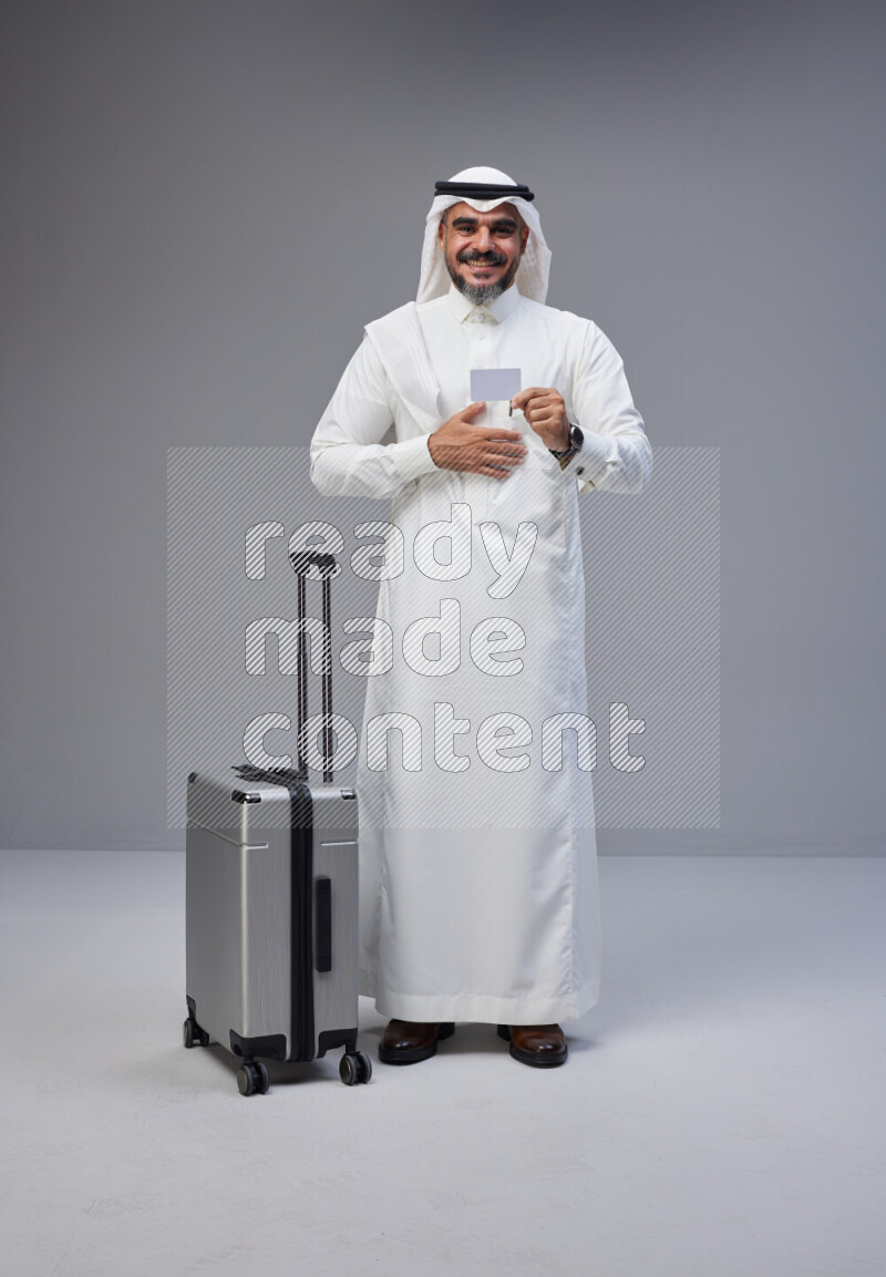 Saudi man wearing Thob and white Shomag standing holding Travel bag and ATM card on Gray background