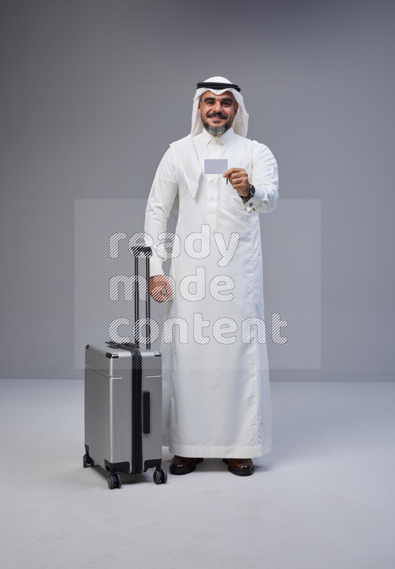 Saudi man wearing Thob and white Shomag standing holding Travel bag and ATM card on Gray background