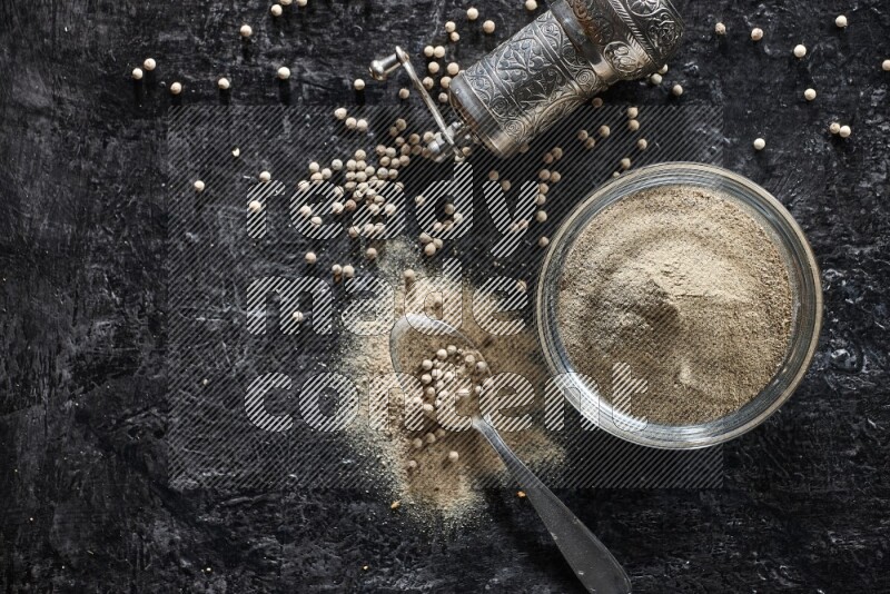 A glass bowl full of white pepper powder with pepper beads, a metal grinder and a metal spoon on textured black flooring