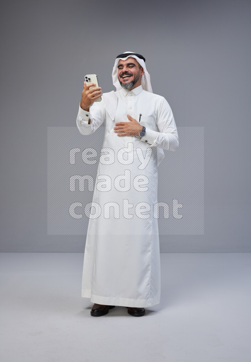 Saudi man Wearing Thob and white Shomag standing texting on phone on Gray background