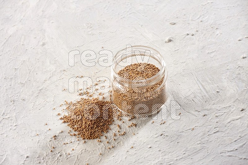 A glass jar full of mustard seeds and more seeds spread on a textured white flooring