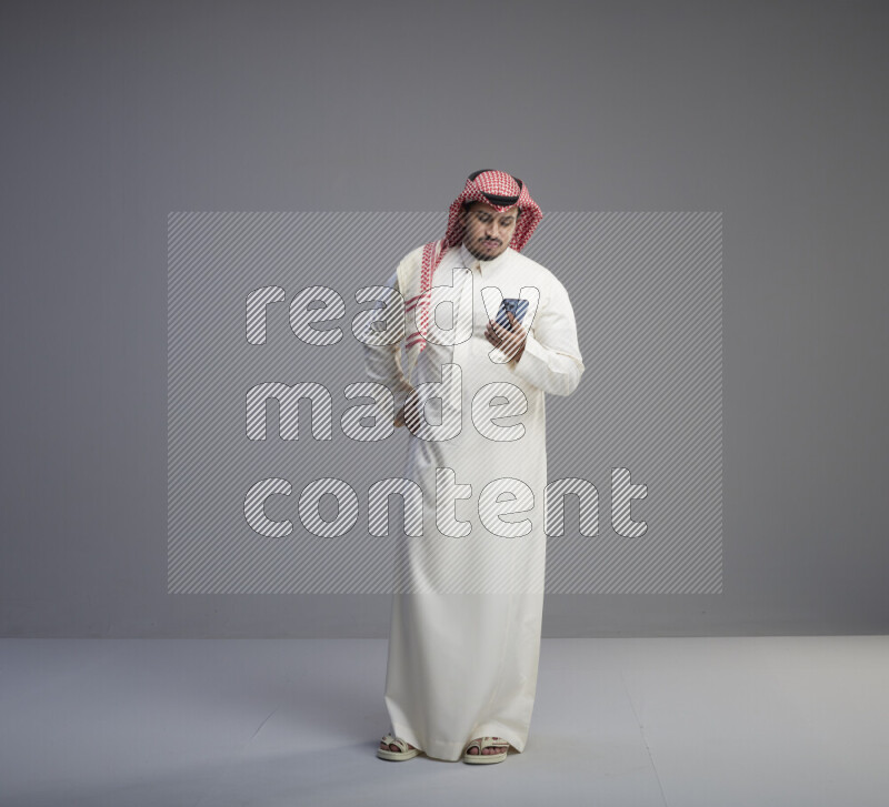 A  Saudi man standing wearing thob and red shomag texting on phone on gray background