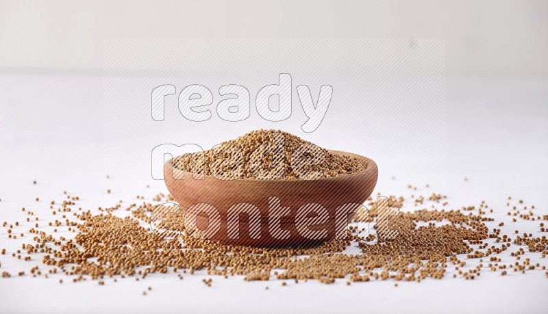 A wooden bowl full of mustard seeds and more seeds spread on a white flooring