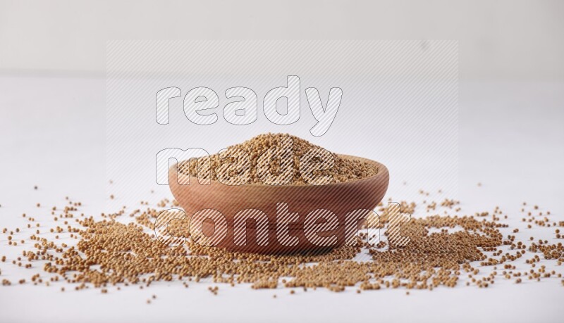 A wooden bowl full of mustard seeds and more seeds spread on a white flooring