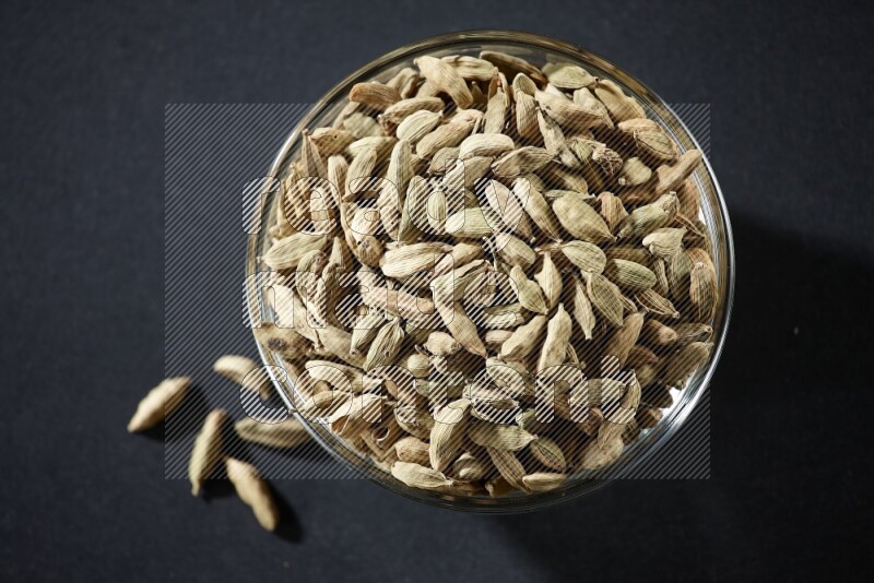 A glass bowl full of cardamom and more seeds spreaded beneath the bowl on black flooring