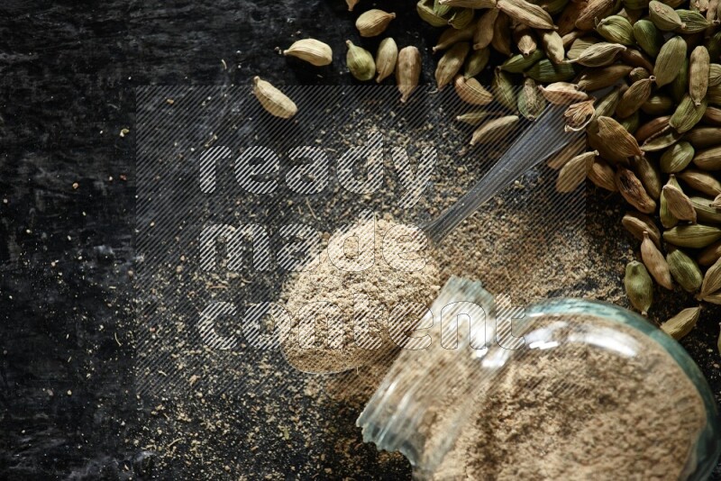 A flipped glass spice jar and a metal spoon full of cardamom powder and cardamom seeds spreaded on textured black flooring