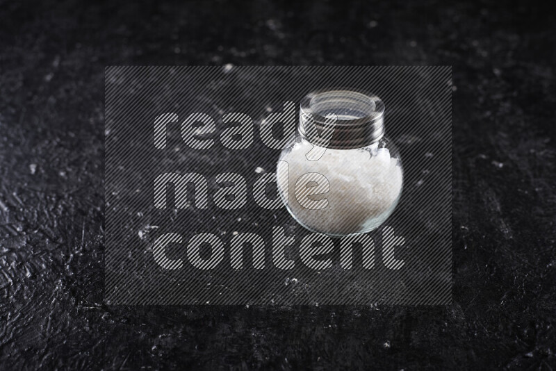 A glass jar full of coarse sea salt crystals on black background