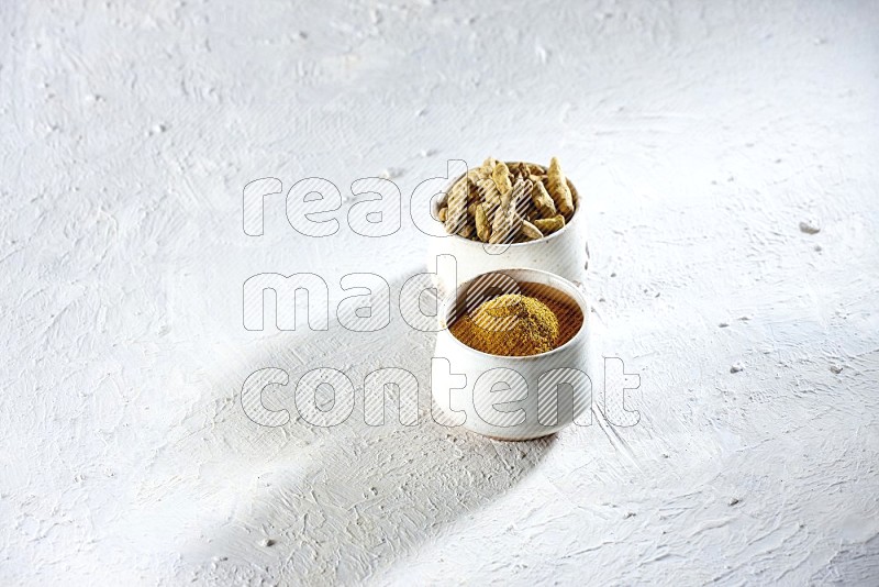 2 beige pottery bowl full of turmeric powder and dried turmeric whole fingers on textured white flooring