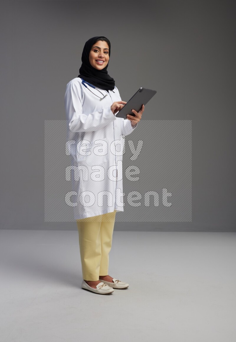 Saudi woman wearing lab coat with stethoscope standing working on tablet on Gray background