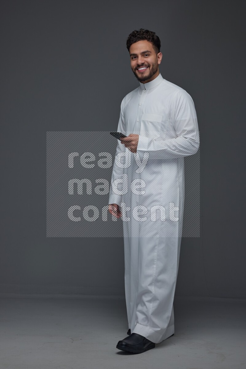 A Saudi man wearing Thobe standing and using his phone eye level on a gray background
