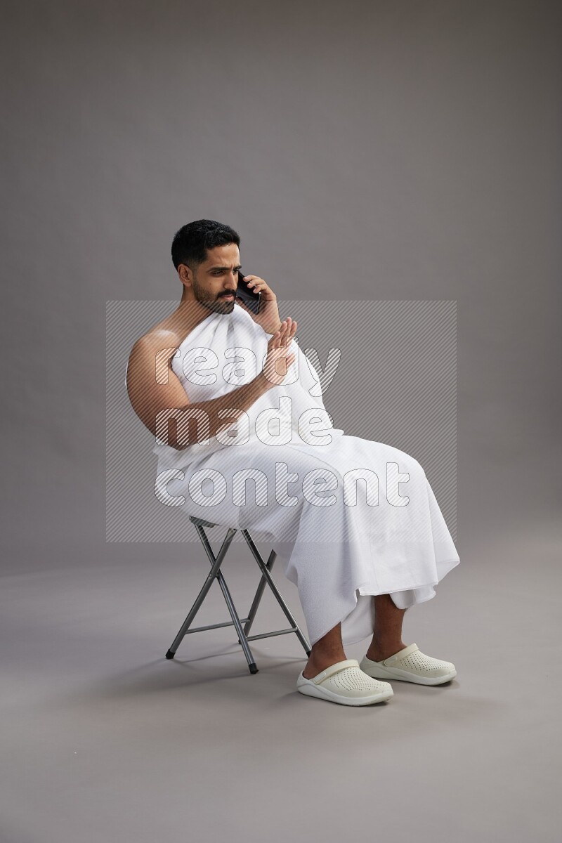 A man wearing Ehram sitting on chair talking on the phone on gray background