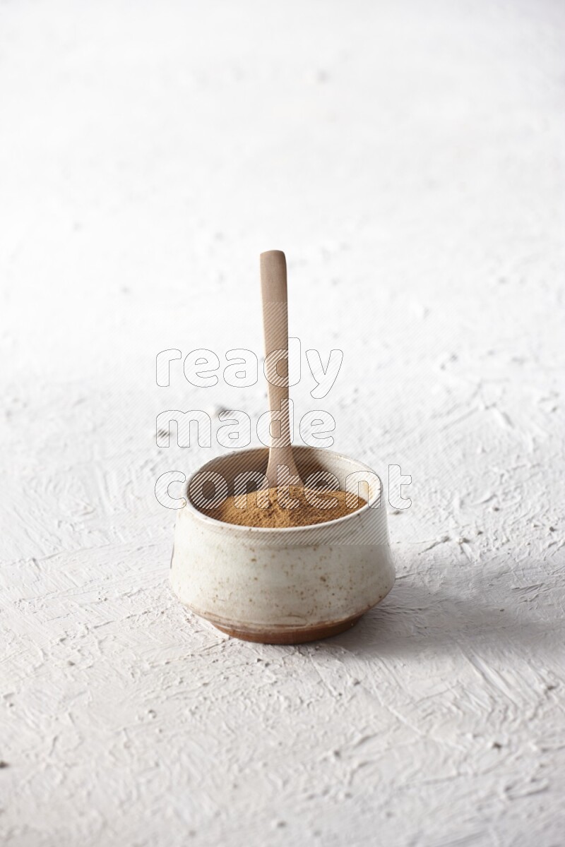 Ceramic beige bowl full of cinnamon powder with a wooden spoon on a textured white background