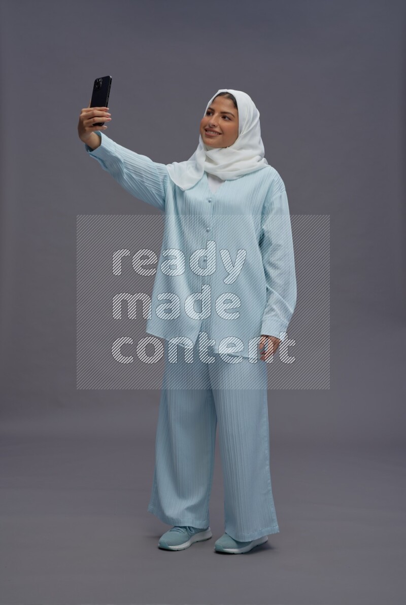 Saudi woman wearing hijab clothes standing taking selfie on gray background