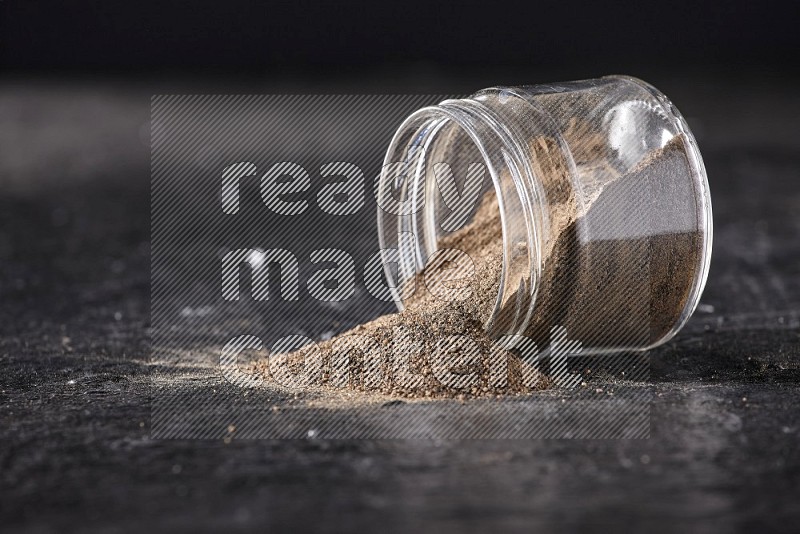 A flipped glass jar full of black pepper powder on a textured black flooring