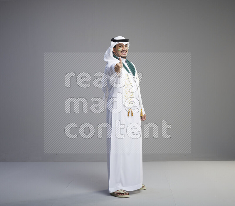 A Saudi man standing wearing thob and white shomag with face painting and Saudi flag scarf on gray background