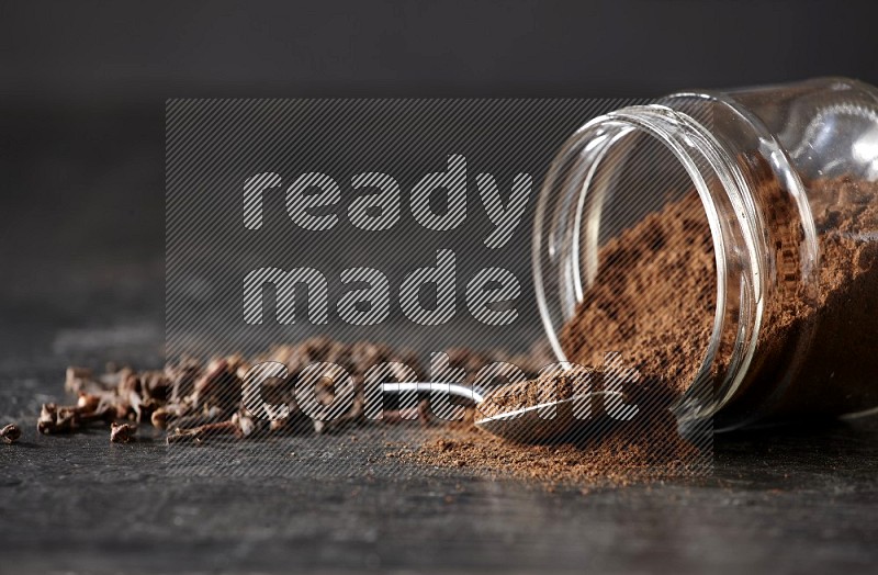 A flipped glass jar and metal spoon full of cloves powder with cloves spread on a textured black flooring