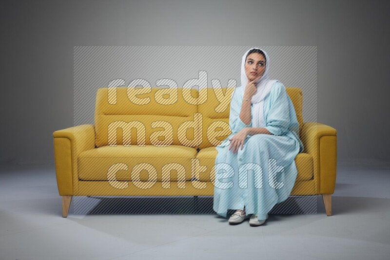 A Saudi woman wearing a light blue Abaya and white head scarf sitting on a yellow sofa and thinking and pointing up eye level on a grey background