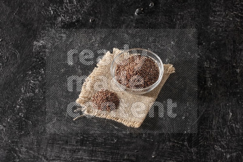 A glass bowl full of flaxseeds with bunch of the seeds on burlap fabric on a textured black flooring