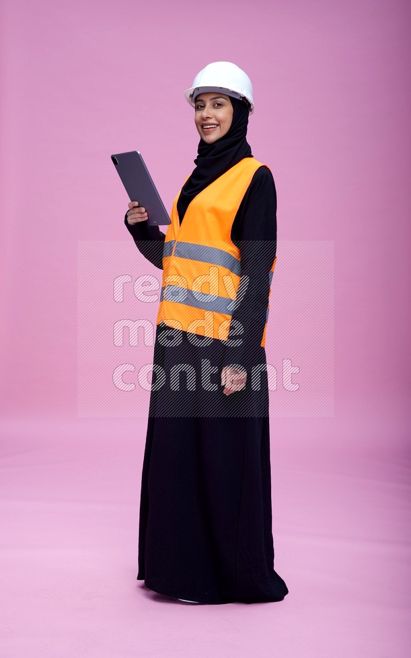 Saudi woman wearing Abaya with engineer vest and helmet standing working on tablet on pink background